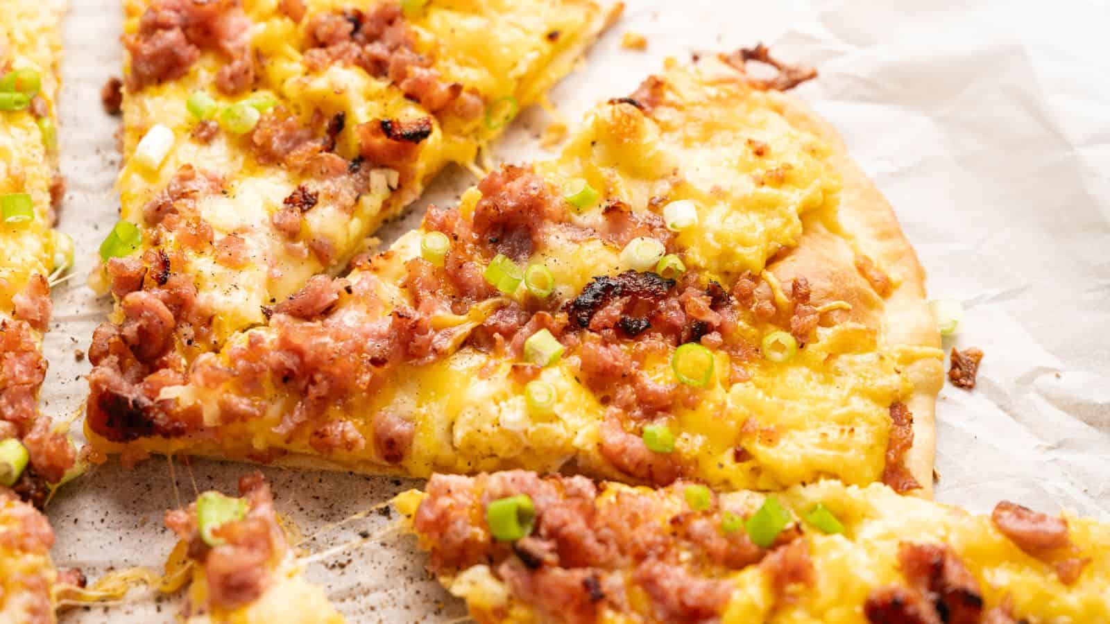 Close-up of a sliced pizza topped with scrambled eggs, bacon pieces, melted cheese, and chopped green onions on parchment paper.