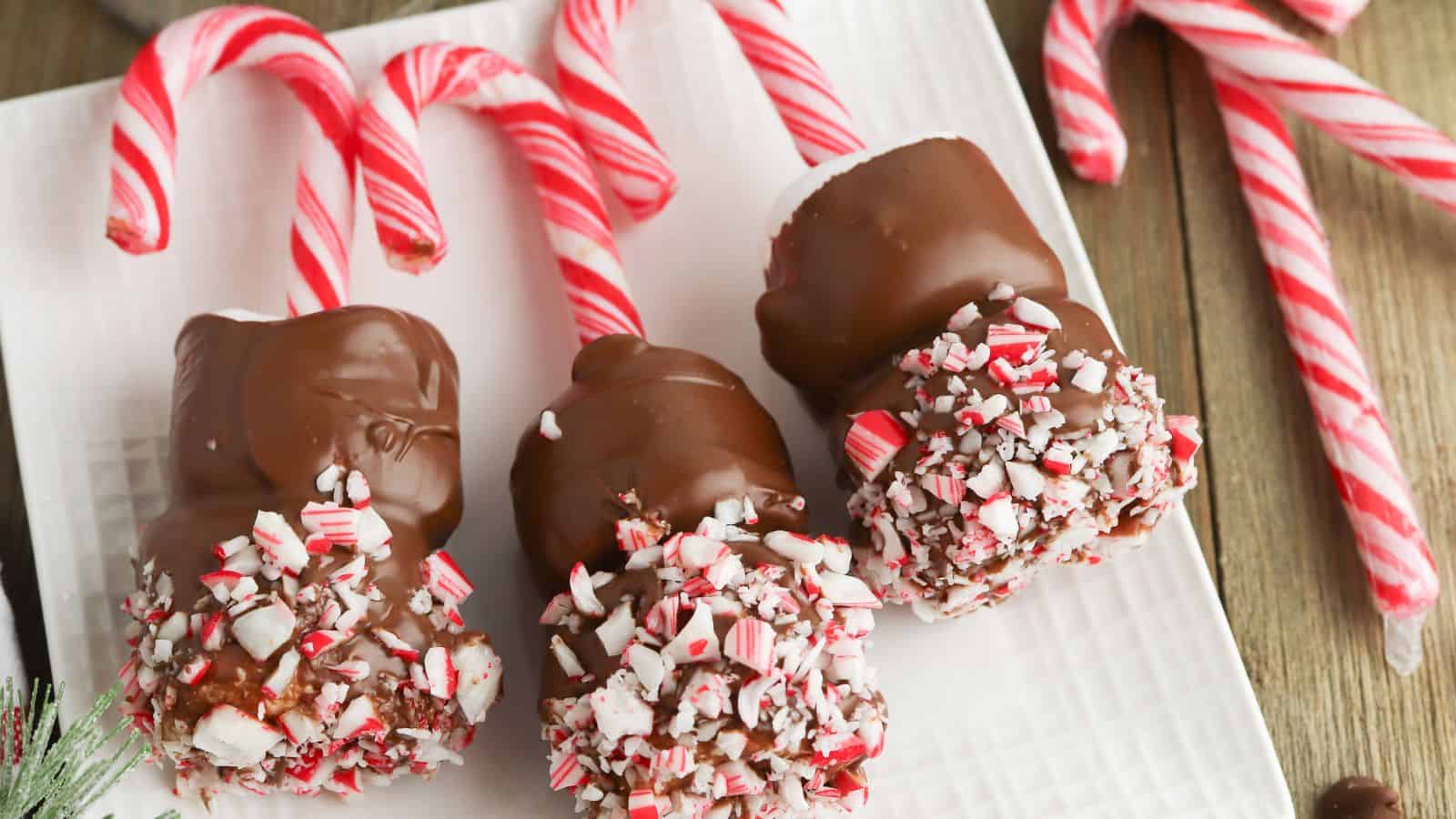 Three marshmallows on candy cane sticks, partially dipped in chocolate and coated with crushed peppermint, are arranged on a white plate.