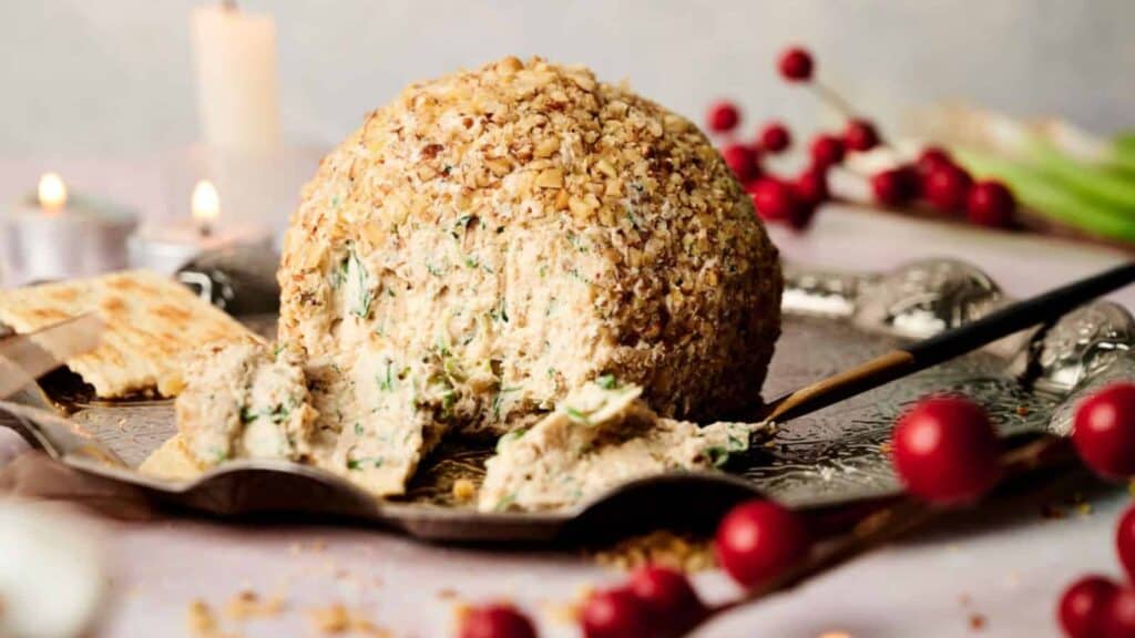A round cheese ball coated in chopped nuts is served on a tray with crackers and festive decorations in the background.