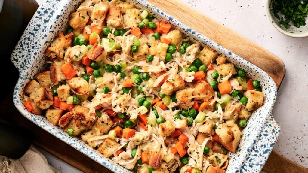 A baked casserole dish filled with cubed bread, shredded chicken, carrots, celery, and green peas on a wooden surface.
