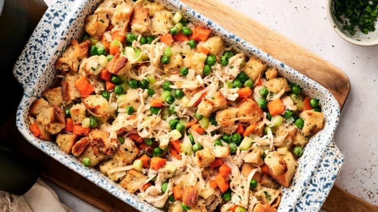 A baked casserole dish filled with cubed bread, shredded chicken, carrots, celery, and green peas on a wooden surface.