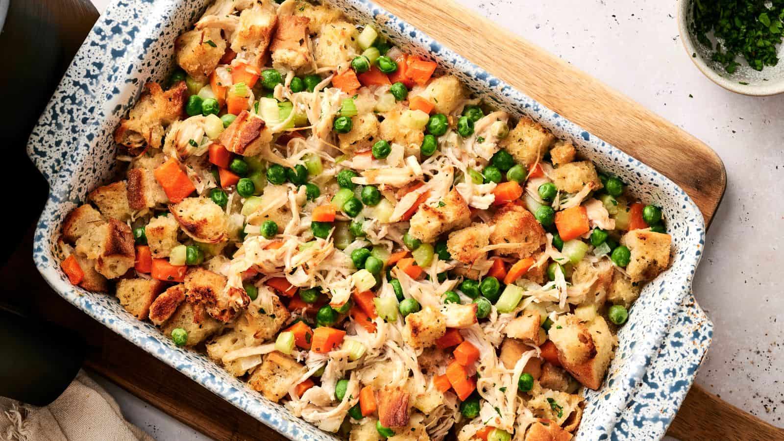 A casserole dish filled with baked bread cubes, shredded chicken, peas, carrots, and celery, arranged on a wooden board.