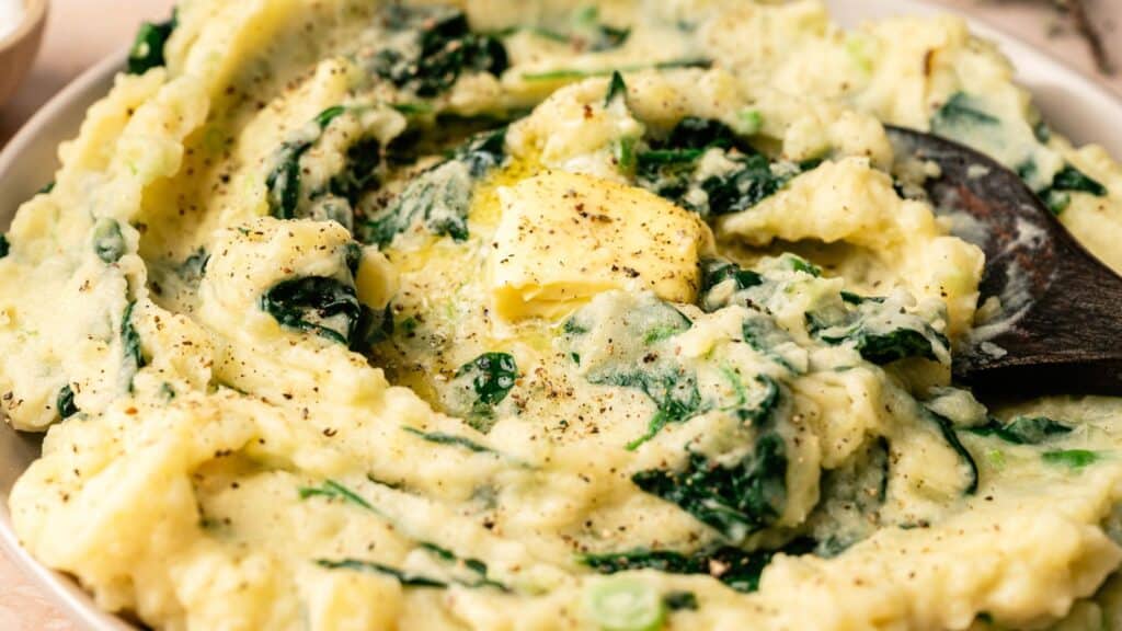 A bowl of mashed potatoes mixed with spinach, topped with a pat of melting butter and black pepper, with a wooden spoon.