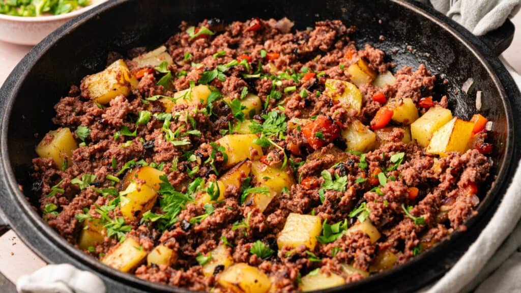 A skillet filled with a ground beef and diced potato hash, garnished with chopped parsley.