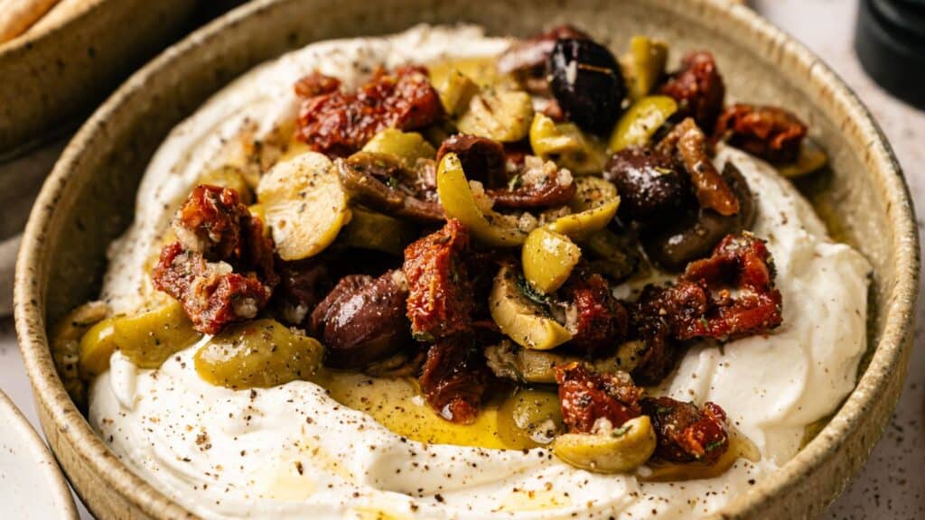 A bowl of creamy whipped feta cheese topped with chopped olives, sun-dried tomatoes, herbs, and a drizzle of olive oil.