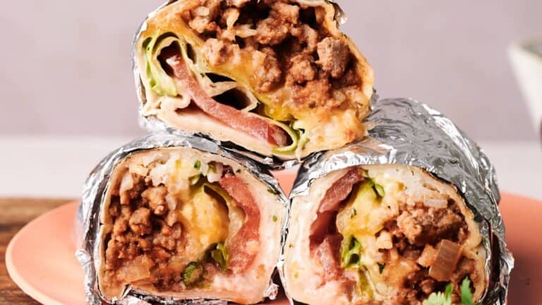 Three halves of beef burritos wrapped in foil are stacked on a pink plate, showing fillings of ground beef, lettuce, tomato, cheese, and sauce.