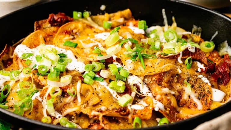 A close-up of a skillet filled with loaded nachos, topped with melted cheese, sliced green onions, sour cream, and various seasonings.