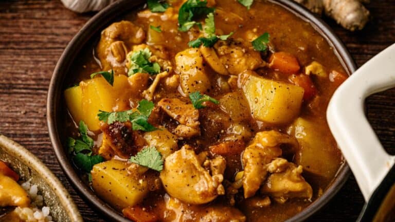 A bowl of chicken curry stew with potatoes, carrots, and fresh cilantro, served on a wooden table.