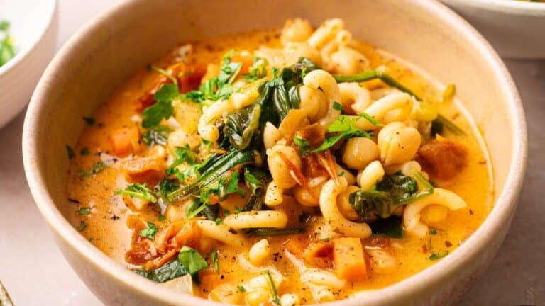 A bowl of pasta soup with chickpeas, leafy greens, diced tomatoes, and herbs in a creamy broth.