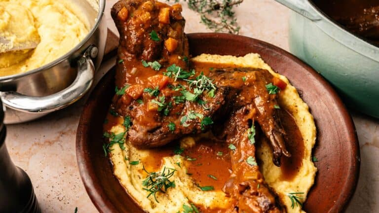 A plate with braised lamb shanks in a tomato-based sauce served over creamy polenta, garnished with fresh herbs.