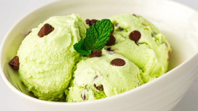 Three scoops of mint chocolate chip ice cream in a white bowl, garnished with a fresh mint leaf.