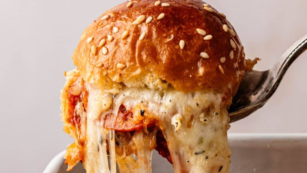 A fork lifts a slider with a sesame seed bun, melted cheese, and tomato sauce, showing a stretch of cheese.