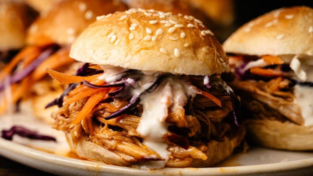 A close-up of a pulled chicken sandwich with carrots, shredded red cabbage, and creamy sauce on a sesame seed bun.
