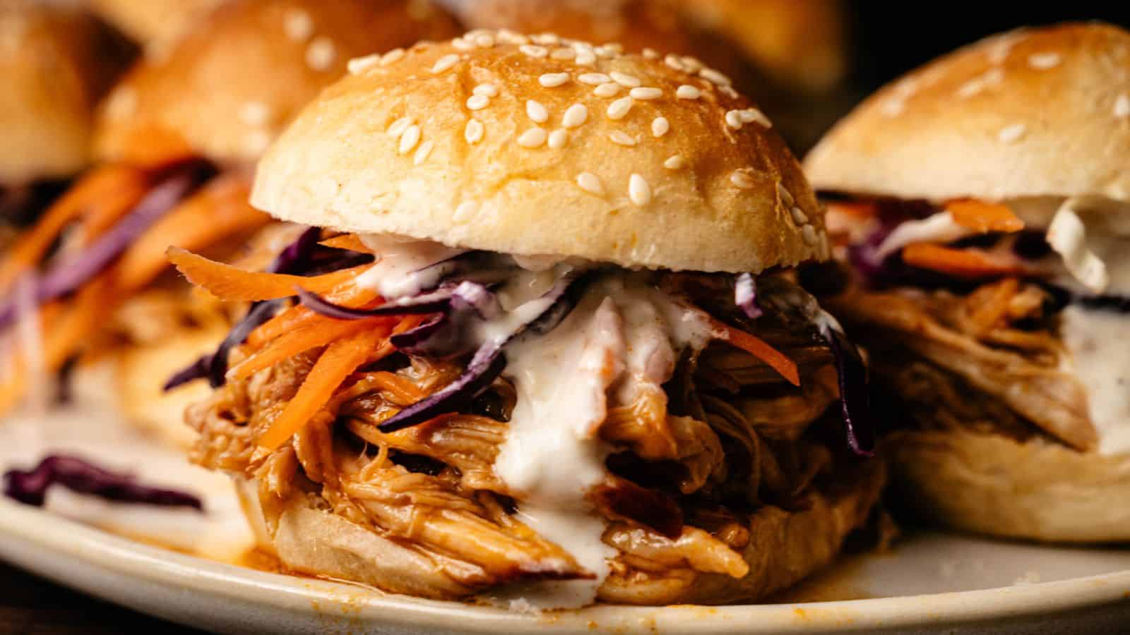 A close-up of a pulled chicken sandwich with sesame seed bun, shredded carrots, cabbage, and creamy sauce.