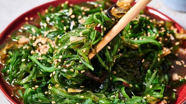 A close-up of chopsticks picking up seasoned seaweed salad topped with sesame seeds from a red plate.