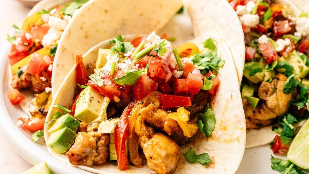 Chicken tacos topped with diced avocado, tomatoes, cilantro, and crumbled cheese served on soft tortillas.