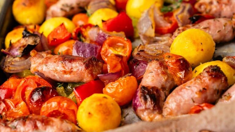 Roasted sausages, cherry tomatoes, red onions, yellow potatoes, and red bell peppers on a baking sheet.