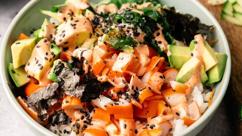 A bowl of rice topped with diced salmon, avocado, carrots, seaweed, black sesame seeds, and a drizzle of creamy orange sauce.