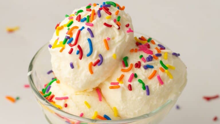 Two scoops of vanilla ice cream in a glass bowl topped with colorful rainbow sprinkles against a plain background.
