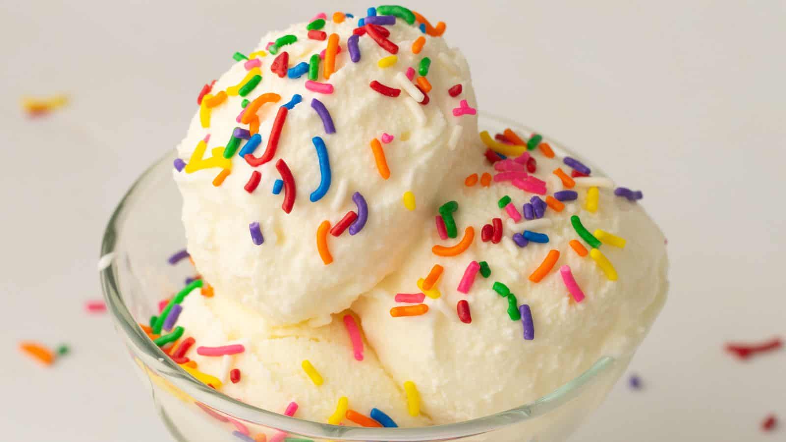 A glass bowl with two scoops of vanilla ice cream topped with colorful rainbow sprinkles, set against a plain white background.