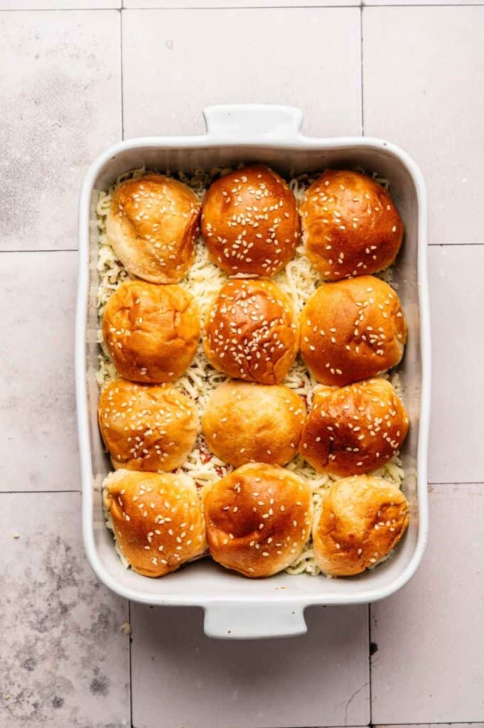 A white baking dish filled with twelve sesame seed slider buns, arranged in rows on top of melted cheese—perfect for serving savory Pizza Sliders.