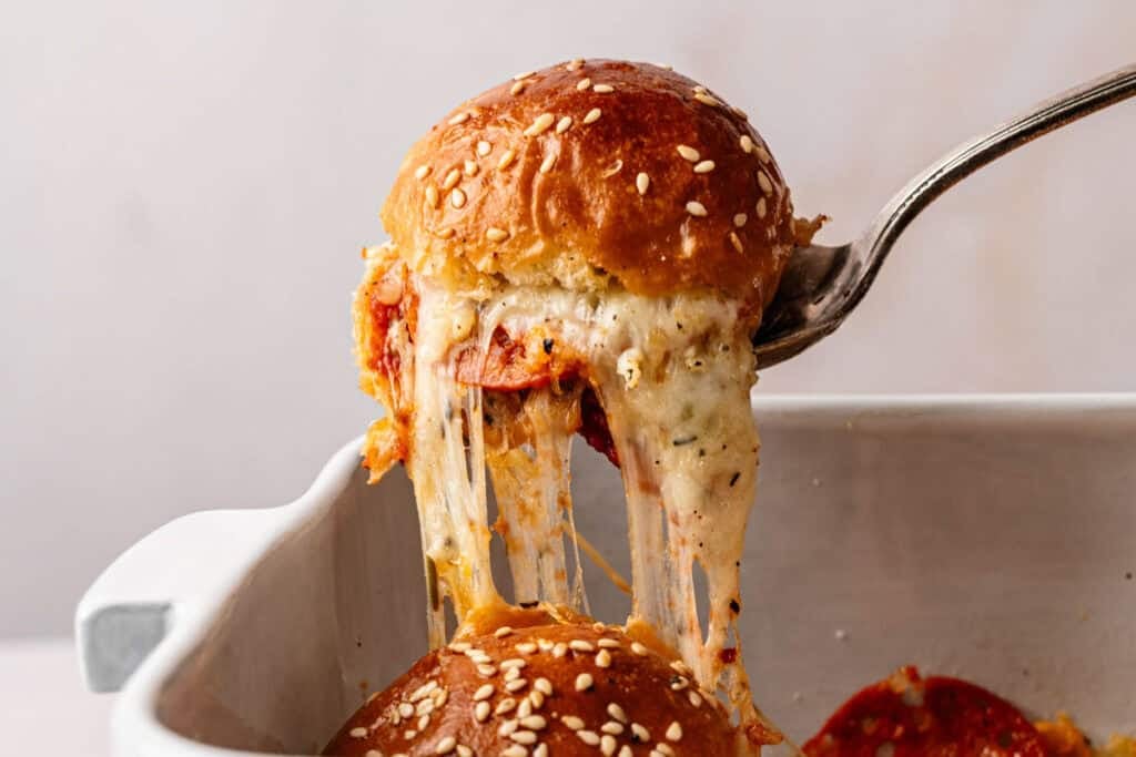 A sesame seed bun filled with melted cheese and pepperoni is lifted from a baking dish, revealing stretchy, gooey cheese—these Pizza Sliders are the ultimate savory snack.