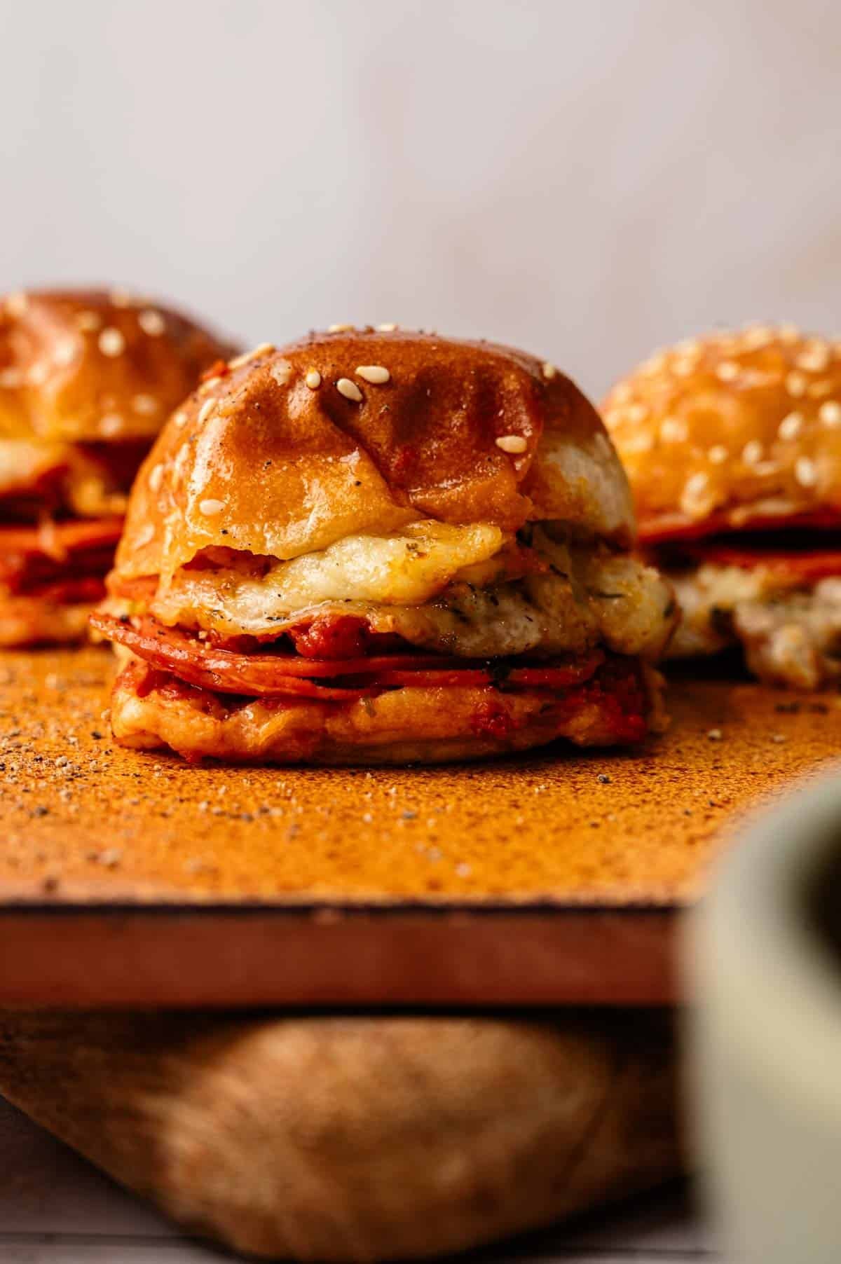 A close-up of Pizza Sliders featuring melted cheese and sliced meat on a sesame seed bun, placed on a wooden surface.
