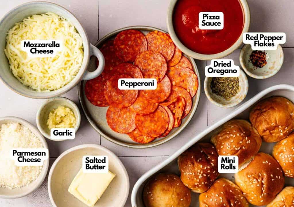 An overhead view of Pizza Sliders ingredients, featuring mozzarella, pepperoni, pizza sauce, mini rolls, parmesan, salted butter, garlic, red pepper flakes, and dried oregano.