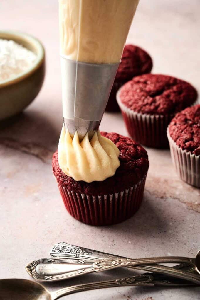 A piping bag is applying cream-colored frosting onto an Air Fryer Red Velvet Cupcake. Several unfrosted cupcakes and a bowl of white ingredients are in the background.