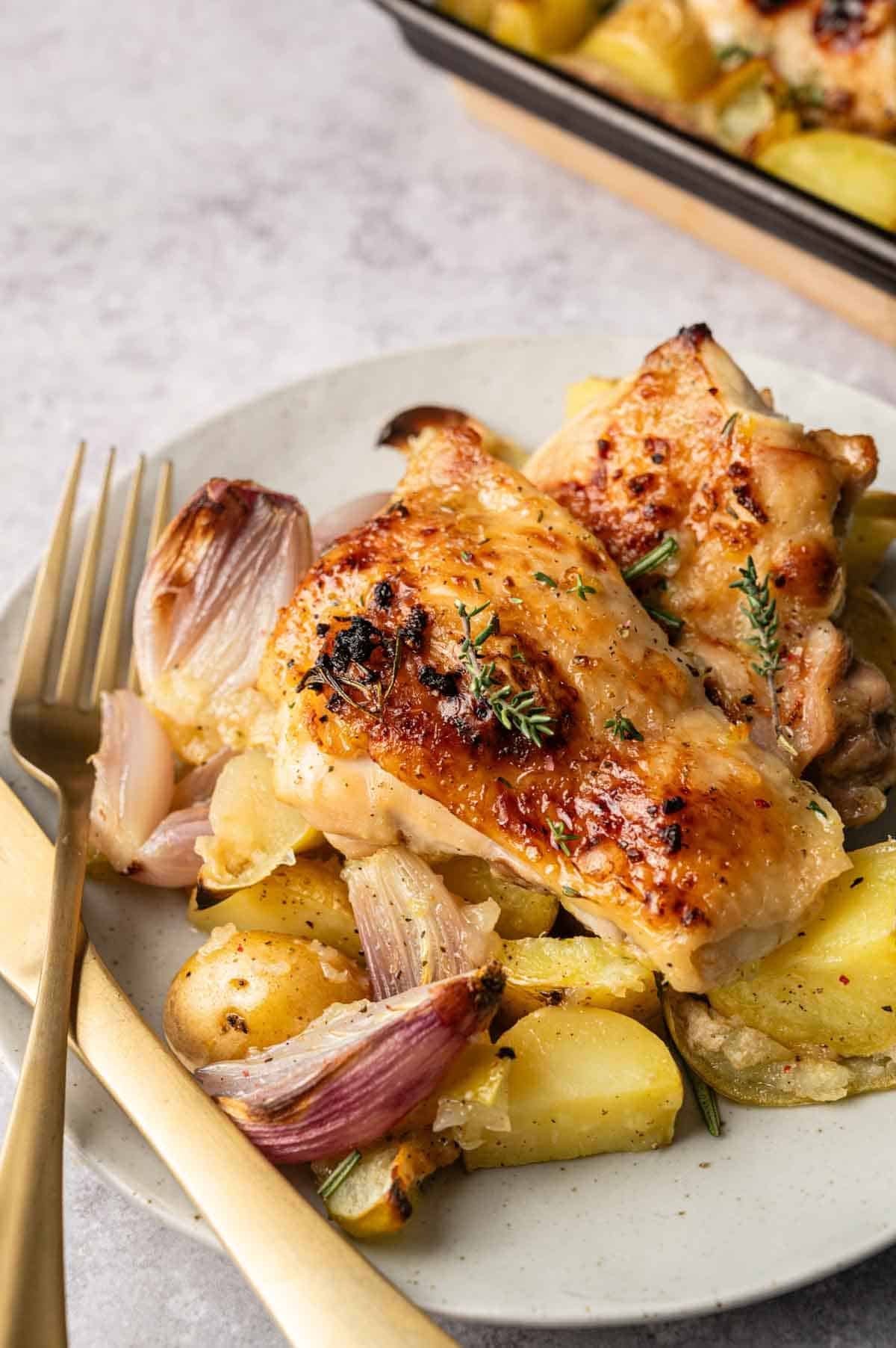 A plate with flavorful Sheet Pan Chicken thighs, potatoes, red onions, and sprigs of thyme, served with a gold fork and knife.