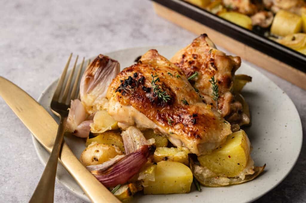 A plate of Sheet Pan Chicken with crispy skin served atop roasted potatoes and onions, garnished with fresh thyme. A gold-colored fork and knife are placed beside the plate.