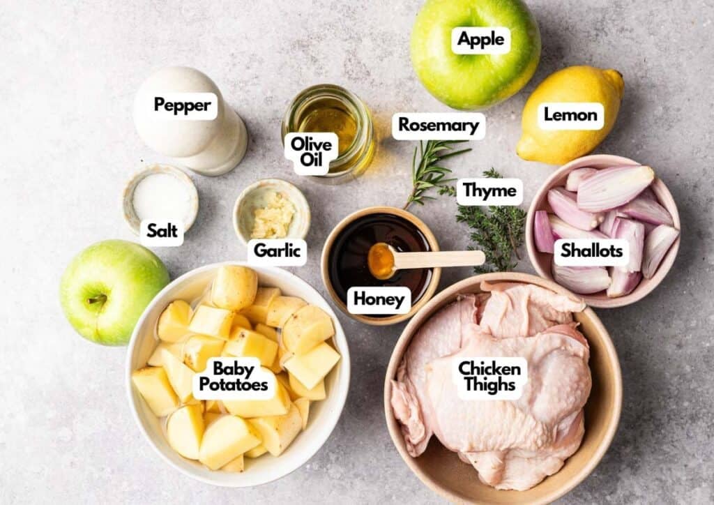 Sheet Pan Chicken ingredients labeled on a countertop: chicken thighs, baby potatoes, apple, shallots, garlic, lemon, olive oil, honey, rosemary, thyme, salt, and pepper.