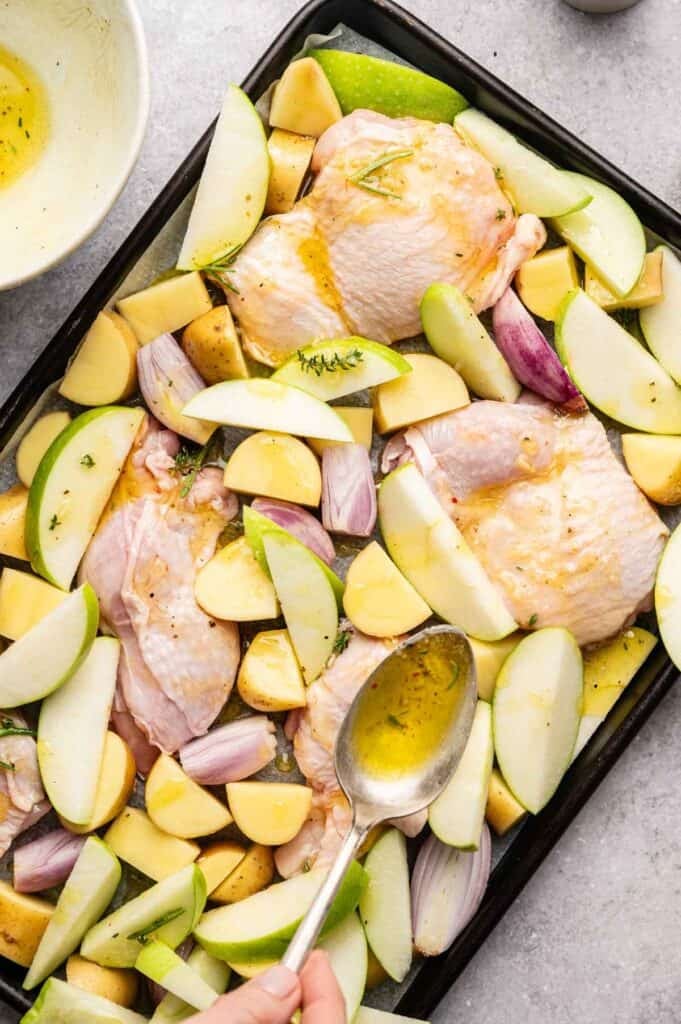 Sheet Pan Chicken legs, chopped potatoes, apple slices, and shallots are arranged on a baking sheet. A hand is drizzling oil over the ingredients with a spoon.