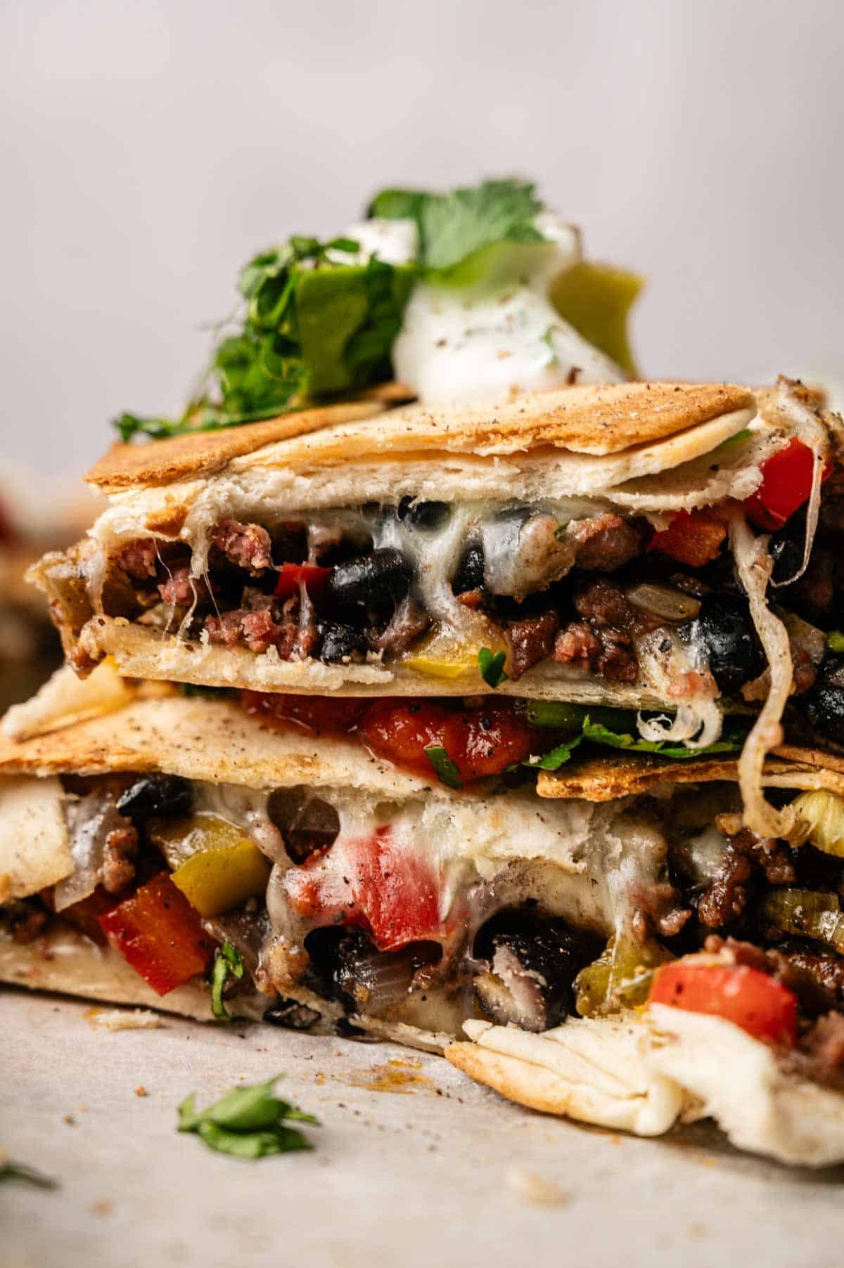 A close-up of stacked Sheet Pan Quesadillas slices filled with ground meat, melted cheese, black beans, and diced vegetables, topped with sour cream and fresh cilantro.