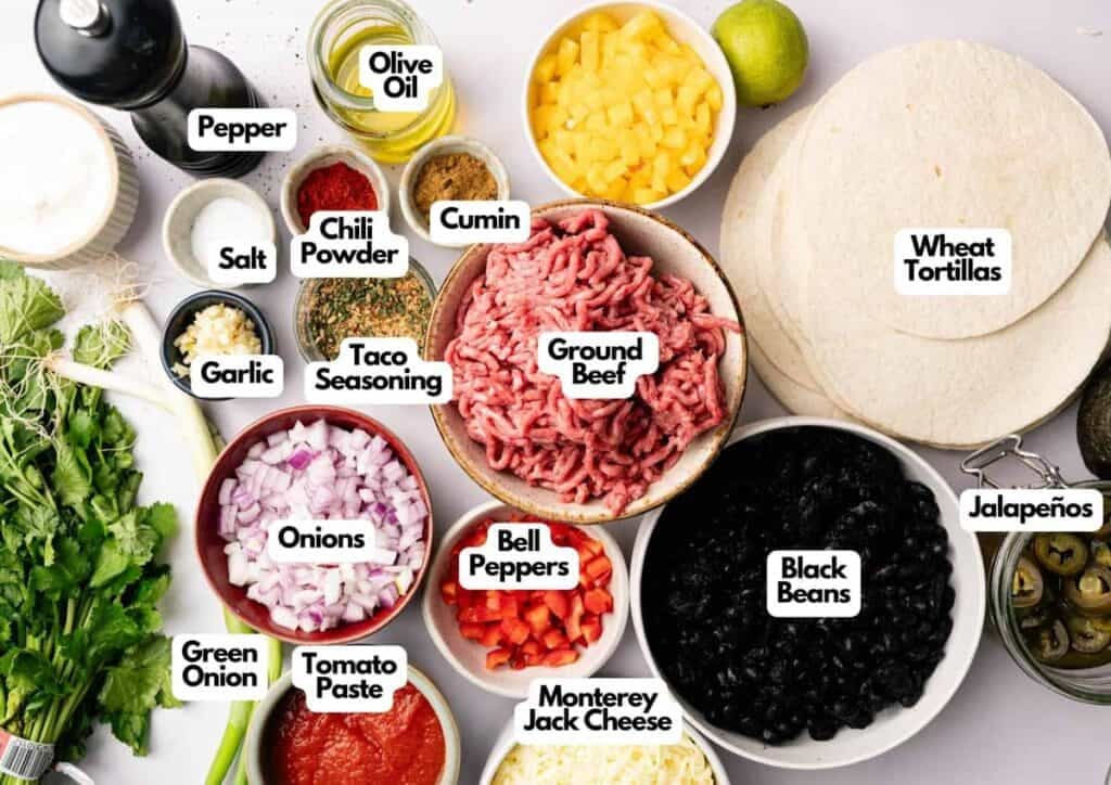 An overhead view of labeled taco ingredients&mdash;perfect for Sheet Pan Quesadillas&mdash;including ground beef, black beans, vegetables, spices, cheese, and wheat tortillas neatly arranged on a white surface.