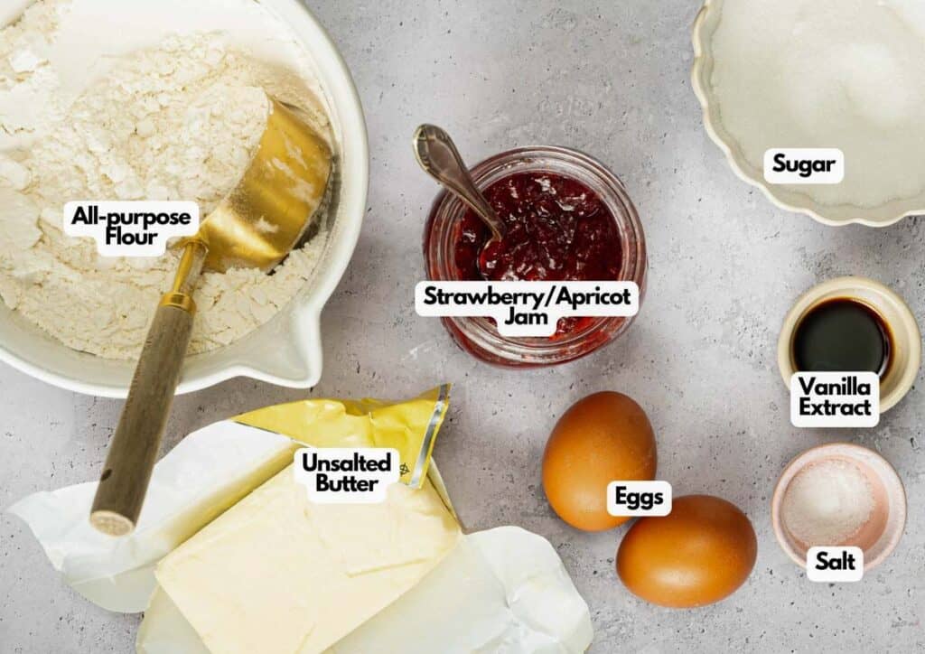 Top-down view of labeled baking ingredients for Air Fryer Thumbprint Cookies: all-purpose flour, unsalted butter, strawberry/apricot jam, eggs, sugar, vanilla extract, and salt on a gray surface.