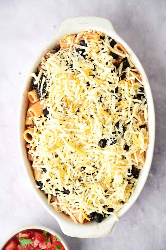 An oval baking dish filled with pasta, black beans, and topped with a generous layer of shredded cheese sits on a light countertop&mdash;perfect for serving as a hearty Walking Taco Casserole.