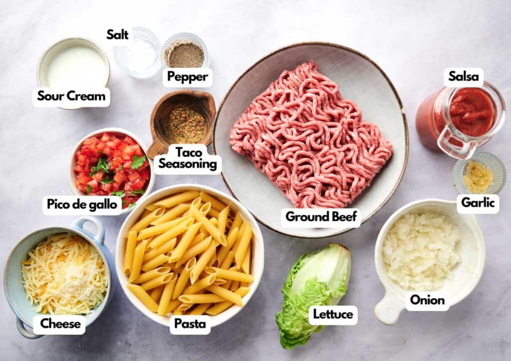 Ingredients for a Walking Taco Casserole pasta dish, including ground beef, pasta, cheese, pico de gallo, lettuce, onion, garlic, salsa, taco seasoning, pepper, salt, and sour cream.