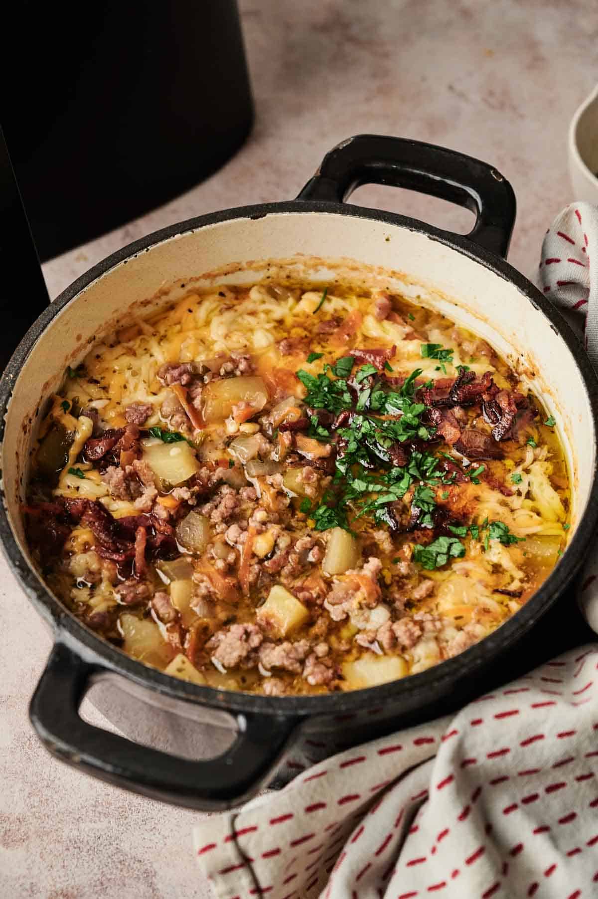 A pot of Air Fryer Cheeseburger Soup with ground meat, potatoes, chopped onions, and fresh herbs, garnished with parsley, sits on a countertop next to a white cloth with red stripes.