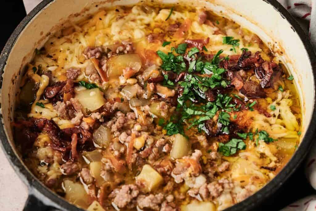 A pot of cooked ground meat, potatoes, cheese, and herbs—reminiscent of Air Fryer Cheeseburger Soup—garnished with chopped parsley and sun-dried tomatoes.