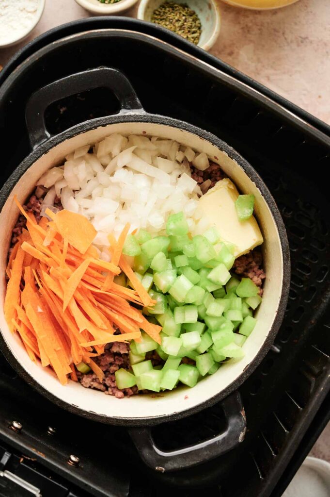 A pot with chopped onions, sliced carrots, diced celery, ground meat, and a chunk of butter is placed inside a black appliance, ready to create a comforting batch of Air Fryer Cheeseburger Soup.