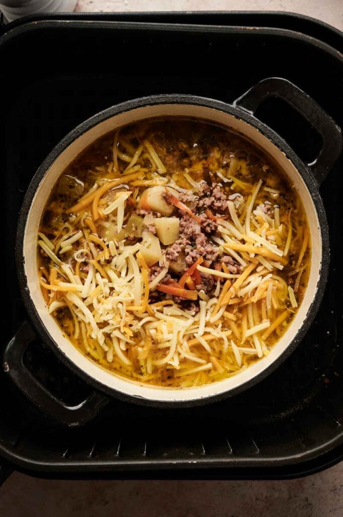 A pot of Air Fryer Cheeseburger Soup, filled with ground meat, chopped potatoes, shredded cheese, and vegetables, sits on a black stovetop.