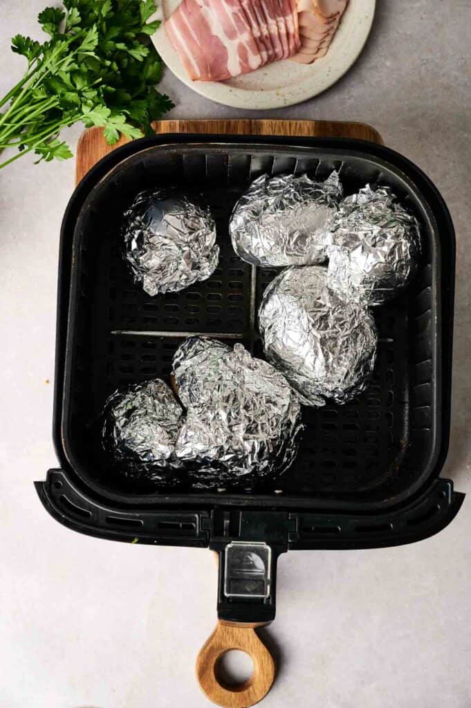 Six foil-wrapped items are placed in an air fryer basket, perfect for prepping Air Fryer Potato Soup. Nearby, a plate with sliced bacon and a bunch of fresh parsley adds fresh flavor and garnishes.