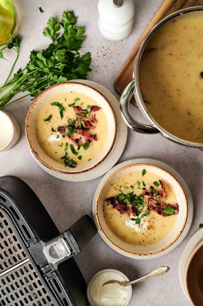 Two bowls of creamy Air Fryer Potato Soup topped with crispy bacon and fresh herbs sit on a counter near a steaming pot of soup, vibrant parsley, an air fryer, and a small bowl of sour cream.