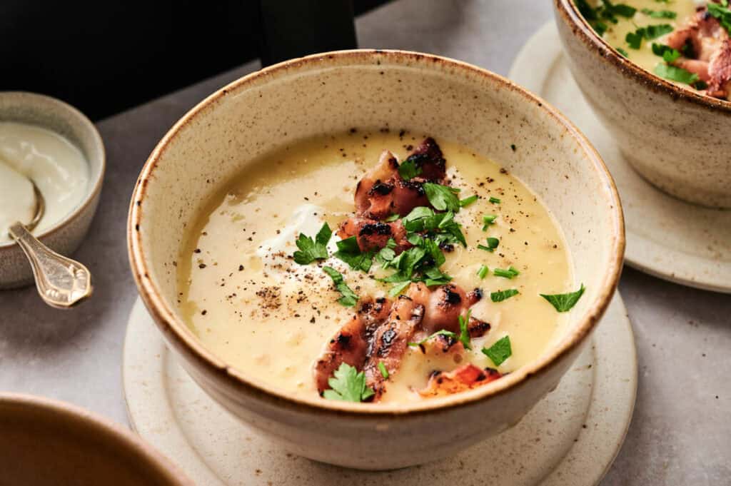 A bowl of creamy Air Fryer Potato Soup topped with grilled bacon pieces, chopped parsley, and black pepper, served on a ceramic plate with a spoon and a small dish nearby.