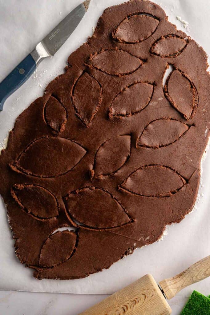 Rolled out chocolate dough with leaf-shaped and American football-shaped cookies cut out, a knife on the side, and a wooden rolling pin partially visible at the bottom.