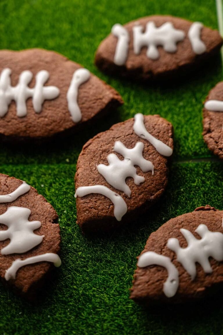 American Football-Shaped Cookies with white icing details are arranged on a green textured surface resembling grass, perfect for sports-themed gatherings.