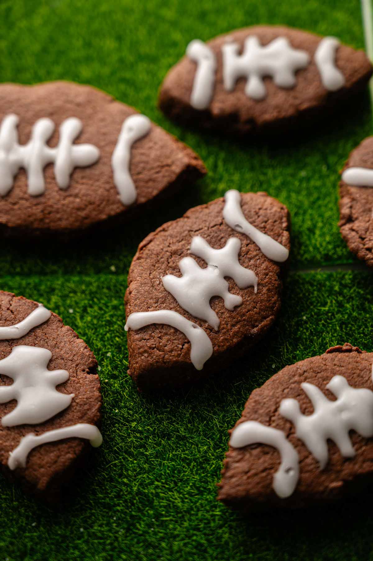 American Football-Shaped Cookies with white icing details are arranged on a green textured surface resembling grass, perfect for sports-themed gatherings.