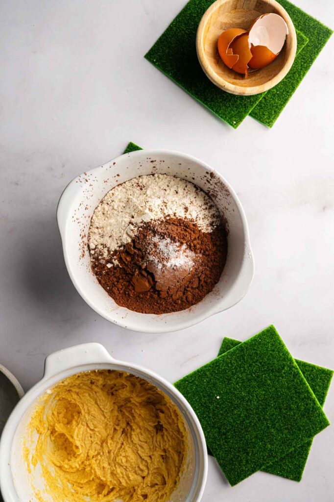 Cocoa powder, flour, and baking powder combined in a white bowl next to creamed butter mixture