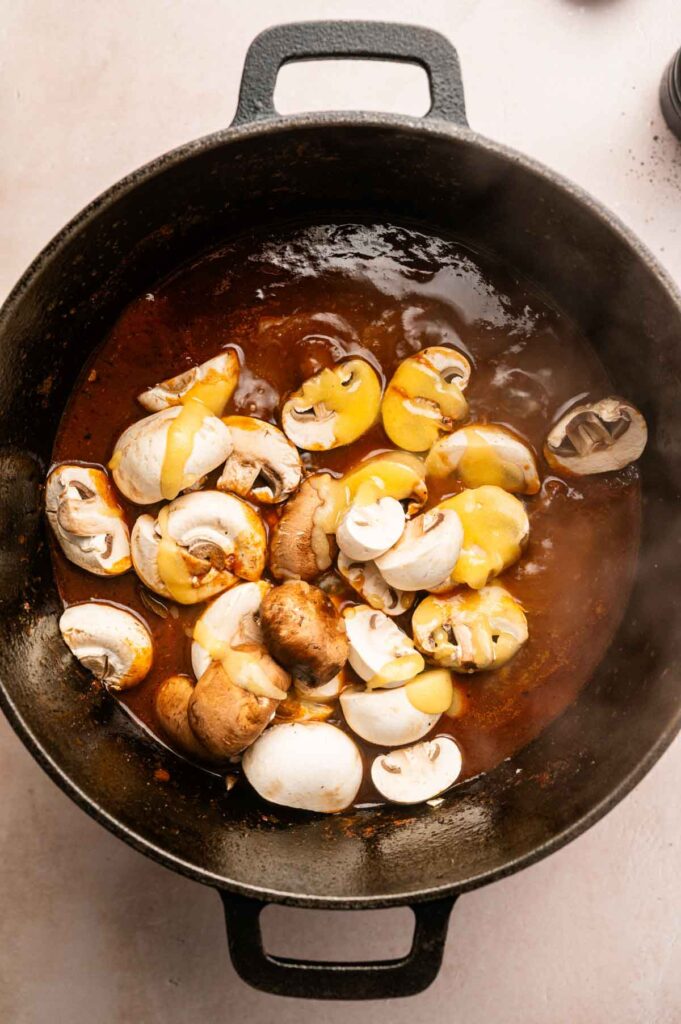 Sliced mushrooms and sauce being cooked in a black cast iron pot as part of a hearty Beef and Guinness Stew, with steam rising from the hot mixture.